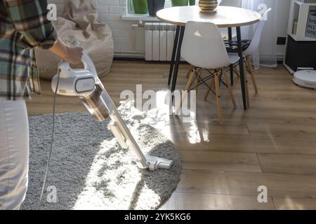 A woman vacuums a round carpet in a house among house plants with a hand vacuum cleaner. General cleaning of the house, cleaning service and housewife Stock Photo