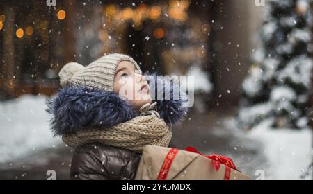Happy girl with a gift box for Christmas catches snowflakes with his tongue on a city in winter with snow on festive market with decorations and light Stock Photo