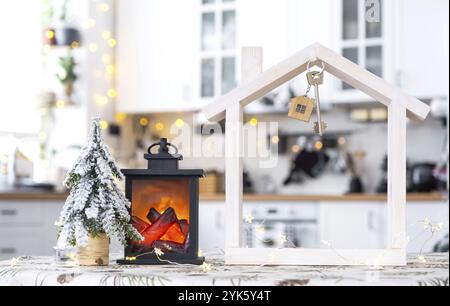 Key and tiny house of a small size on cozy home with Christmas decor on table of festive white kitchen. Gift for New Year. Insurance, moving to new ho Stock Photo