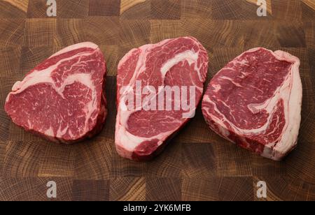 Close up three raw marbled ribeye beef steaks over end grain cutting board of wooden butcher block, high angle view, personal perspective Stock Photo