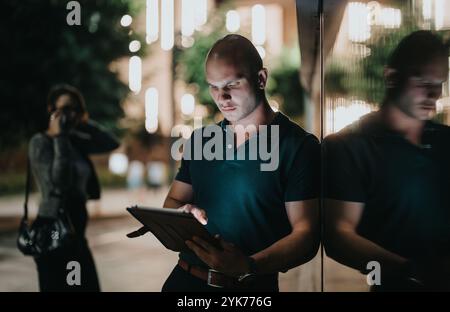 Businessman using tablet at night outdoors reflecting on glass wall with pedestrians in background Stock Photo