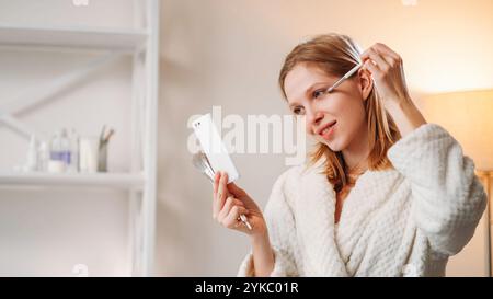 Beauty care. Female routine. Pleasant smiling lady applying makeup using powder brush looking in mirror enjoying her reflection in white robe while holding cosmetic tools in hand. Stock Photo