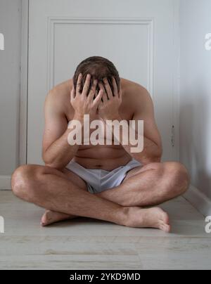 Caucasian man sitting on the floor, dressed in white shorts, with his legs crossed and hands covering his face.  Concept for headache or mental health Stock Photo