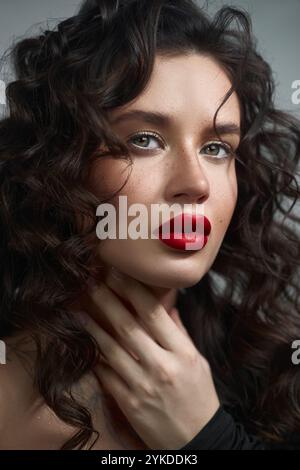 woman with beautiful curly hair poses elegantly, accentuating her striking features with bold red lips in a soft-lit environment, creating an enchanti Stock Photo
