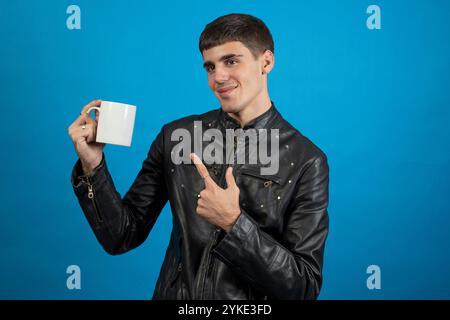 Young man wearing black leather jacket holding and pointing at white mug on blue background Stock Photo