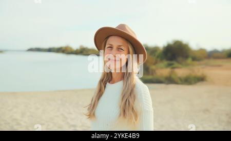 Stylish happy woman standing on sunny beach, warm weather, smiles, wear hat, white sweater, on sea coast background Stock Photo