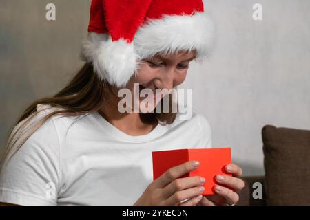 Young woman in a Santa Claus hat opens a small red Christmas gift box and smiles, anticipating the surprise inside Stock Photo