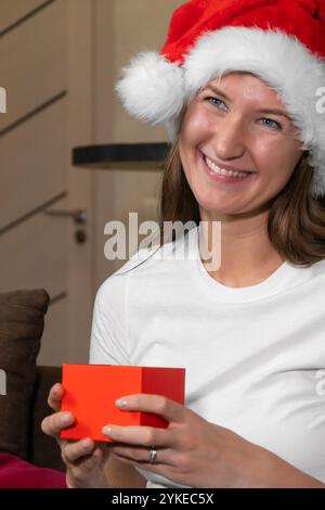 Woman in a santa hat smiles happily, holding a red christmas gift in a cozy home setting, creating a festive and joyful atmosphere. Vertical photo Stock Photo