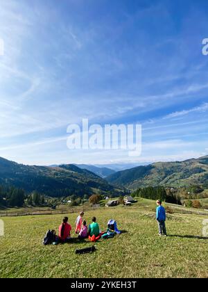 A family sits on a grassy hill enjoying a picnic, surrounded by picturesque mountains and a vibrant blue sky. The scene captures the essence of nature Stock Photo