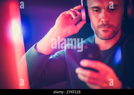 A man enjoys music using headphones while checking his smartphone in a dark setting, illuminated by colorful lights. Stock Photo