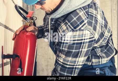 A worker dressed in plaid shirt inspects a fire extinguisher mounted on a wall, ensuring safety compliance in a commercial setting. Stock Photo