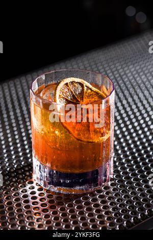 A classic whiskey old fashioned served with a slice of dried citrus in a clear glass The cocktail rests on a textured metal surface, highlighting its Stock Photo