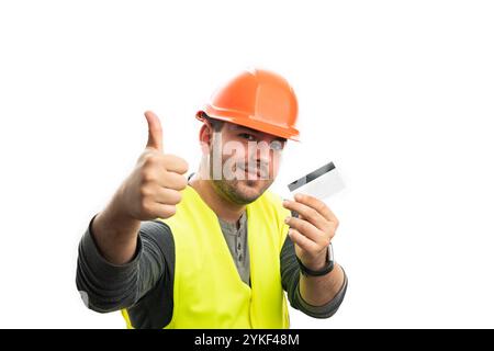 Friendly male constructor smiling waring orange hardhat and safety vest showing thumb up like gesture holding credit or debit bank card isolated on wh Stock Photo