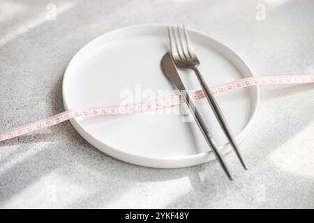 From above an empty white plate, fork, and knife wrapped with a pink measuring tape on a textured grey background, symbolizing portion control and wei Stock Photo