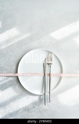 Top view of a dieting concept featuring a white plate, a fork, and a measuring tape on a textured grey background, highlighting portion control and we Stock Photo