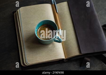 Top view of a coffee mug on an open notepad alongside a pen and leather cover, depicting an organized workspace. Stock Photo