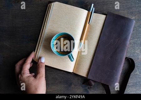 Top view of a cropped unrecognizable woman hand on a wooden desk with a notepad, pen, and coffee cup, illustrating a focused work setting. Stock Photo