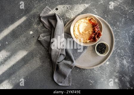 Top view, cropped unrecognizable shot of hummus topped with Jamon, served on a gray ceramic plate with a linen napkin and black sesame seeds on a rust Stock Photo