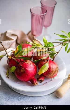 Vibrant ripe pomegranates, with attached twigs and green leaves, set on a white marble plate. The image also features soft pink glasses and a beige na Stock Photo