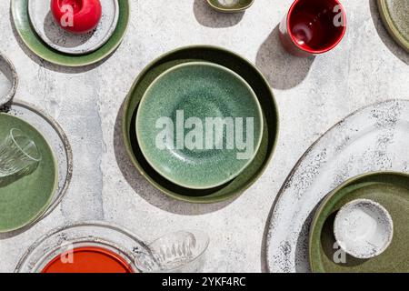 Top view of a vibrant collection of ceramic ware including green and white bowls, plates, and a red candle, artistically arranged on a textured gray s Stock Photo