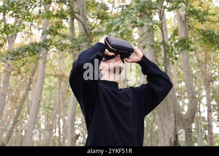 A young trans man dressed in a black sweater uses a virtual reality headset among tall trees in a serene forest, embracing advanced technology in a na Stock Photo