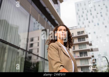 A young Brazilian businesswoman stands confidently in a modern urban environment, dressed in a stylish plaid blazer, gazing forward with determination Stock Photo