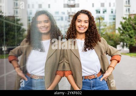 A cheerful Brazilian woman in business attire poses confidently with her reflection in a glass pane in an urban landscape, epitomizing modern, empower Stock Photo