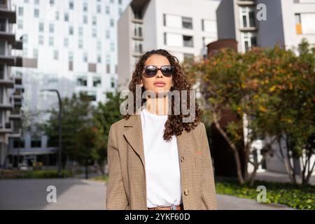 An elegant Brazilian businesswoman wearing sunglasses and a jacket stands confidently in an urban setting with modern buildings in the background Stock Photo