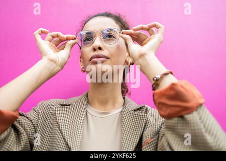 A young Brazilian businesswoman in stylish eyewear and a chic blazer, adjusting her glasses, poses confidently against a vivid pink background Stock Photo