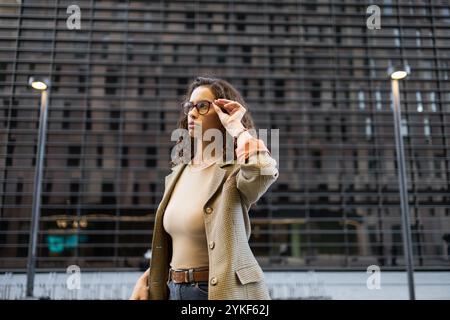 A young Brazilian businesswoman corrects her glasses against an urban backdrop, exuding confidence and professionalism in a stylish blazer Stock Photo