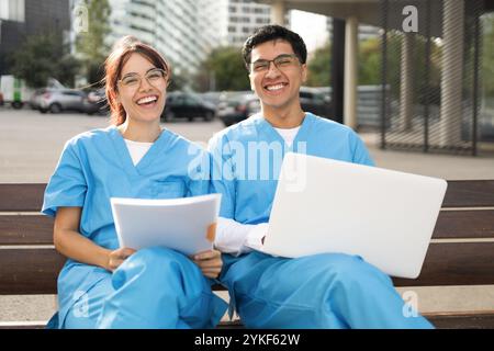 Two smiling nursing students, one male and one female, sit together on a bench outside, studying from papers and a laptop in their blue scrubs Stock Photo