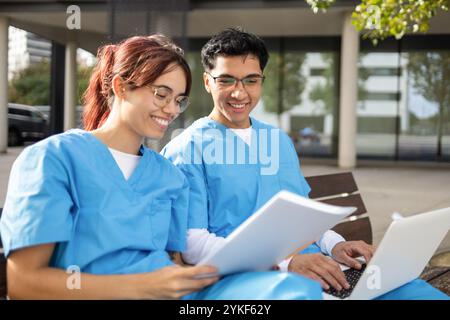 Two smiling nursing students, one male and one female, sit together on a bench outside, studying from papers and a laptop in their blue scrubs Stock Photo