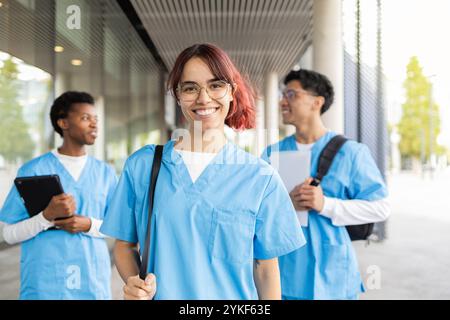 A young female nursing student with pink hair and glasses smiles confidently at the camera, flanked by classmates in a modern educational setting Stock Photo
