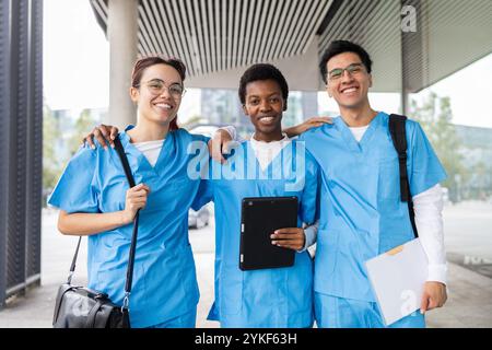 Three happy nursing students in blue scrubs stand together outside their university, sharing a joyful moment Stock Photo