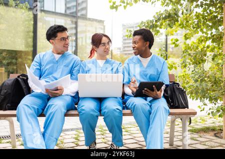 Three nursing students in blue scrubs are engaged in a study session outside, using a laptop and digital tablets, surrounded by lush greenery Stock Photo