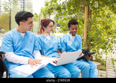 Three nursing students wearing blue scrubs are engaged in studying together using a laptop and notebooks in an outdoor park setting, showcasing a mome Stock Photo