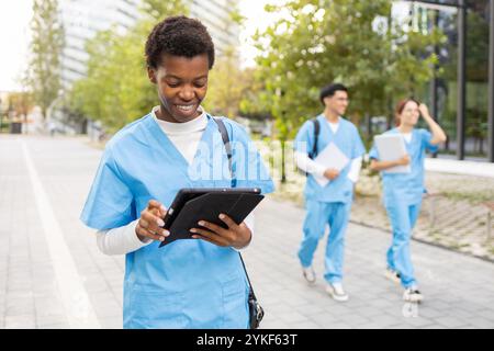 A young female nurse student focuses intently on a digital tablet while walking on a university campus, with her peers in the background also studying Stock Photo