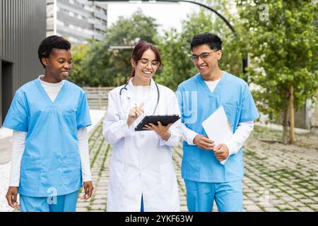 Three young nursing students of diverse backgrounds converse joyfully while using a digital tablet outside their medical campus Stock Photo