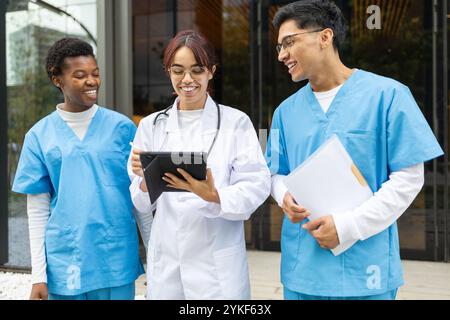 Three multiethnic nurse students, engaged in a discussion hold a tablet and papers outside a modern healthcare facility Stock Photo