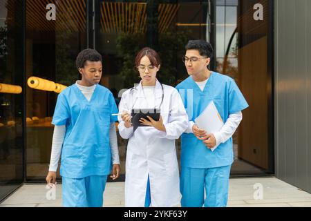 Three diverse nursing students, in medical scrubs, actively engage in a learning discussion using a digital tablet outside a modern university buildin Stock Photo