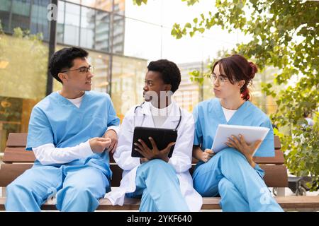 A diverse group of nursing students sits on a bench outdoors, engaged in an educational discussion with tablet and notes in hand, against an urban cam Stock Photo