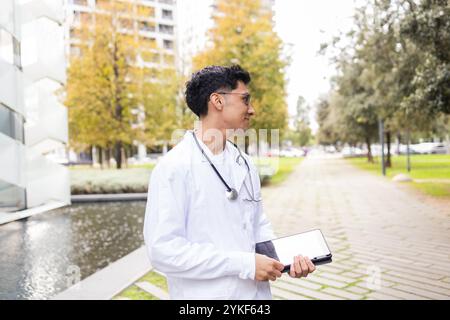 A young male nursing student in a white coat and stethoscope uses a digital tablet while walking outside a modern urban campus Stock Photo