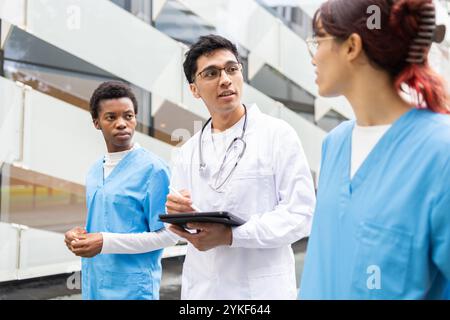 Two nurse students in scrubs engage in a focused discussion with a male doctor holding a clipboard, outside a modern hospital building Stock Photo