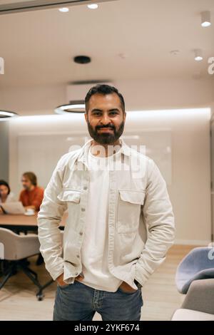 An indian man smiling in a bright, modern coworking space In the background, two colleagues are engaged in a discussion The ambiance is chic and invit Stock Photo