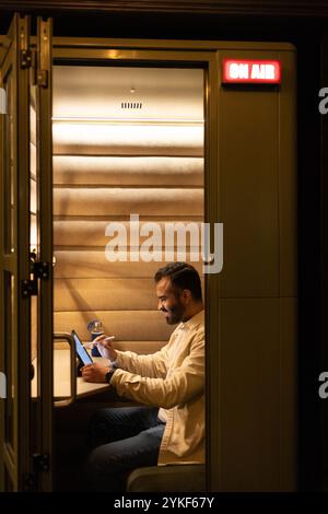 An indian person works attentively on a tablet inside a soundproof booth, illuminated by warm lighting The on air sign adds a professional touch to th Stock Photo