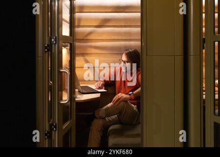A person is comfortably working on a laptop in a cozy, stylish coworking space nook The warm lighting and modern design create an inviting atmosphere Stock Photo