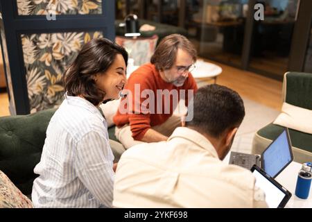 Three colleagues collaborate in a vibrant coworking space A woman smiles, engaging with two colleagues using laptops The creative environment fosters Stock Photo