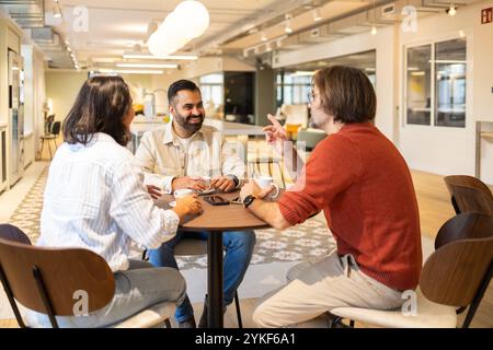 Three professionals share ideas in a lively coworking space, enjoying coffee and conversation The modern setting is bright and open, fostering creativ Stock Photo