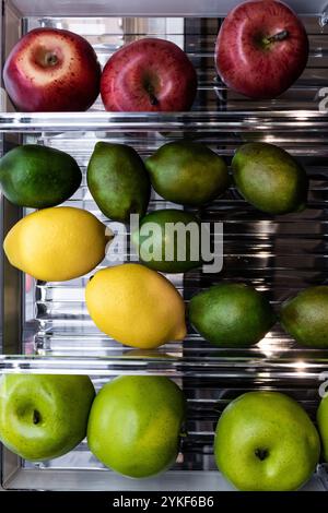 Top view of various fresh fruits neatly arranged on refrigerator shelves, including red and green apples, yellow lemons, and green limes. Stock Photo
