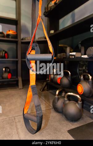 A detailed view of an orange and black suspension trainer in a well-equipped gym, with kettlebells and other fitness gear in the background. Stock Photo